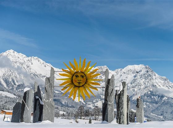 Eine winterliche Landschaft mit schneebedeckten Bergen und einem bunten Sonnensymbol in der Mitte. Rund um das Symbol stehen große Steine, die eine mystische Atmosphäre schaffen.