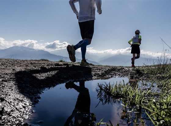 Zwei Läufer joggen auf einem unebenen Weg mit Blick auf die Berge. Im Vordergrund spiegelt sich der Himmel in einer Pfütze.