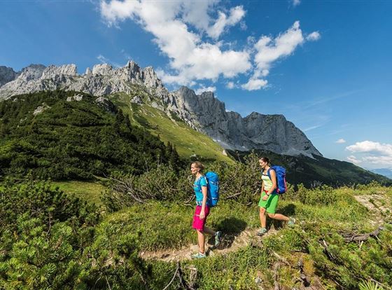 Zwei Wanderer gehen auf einem Pfad in den Bergen. Die Landschaft ist grün mit felsigen Gipfeln und klarem Himmel.