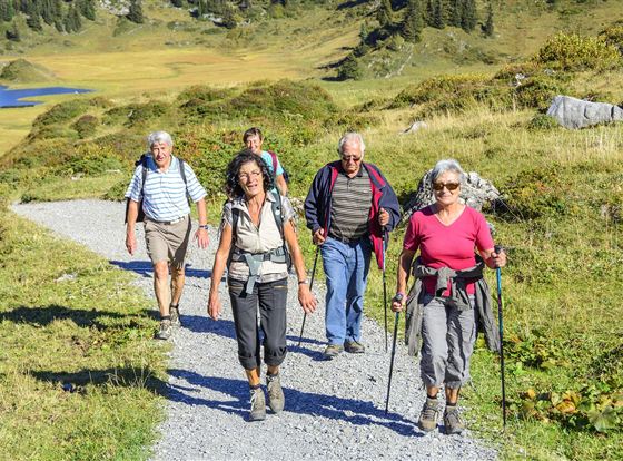 Eine Gruppe von fünf Personen wandert auf einem Schotterweg durch eine malerische Landschaft. Im Hintergrund sind grüne Hügel und ein blauer See zu sehen.