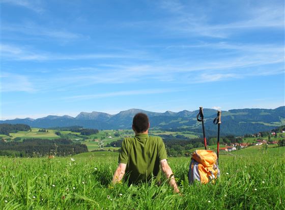 Ein Wanderer sitzt auf einer grünen Wiese und schaut auf die Berge. Der Himmel ist klar und blau, im Hintergrund sind sanfte Hügel und ein kleines Dorf zu sehen.