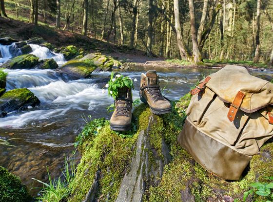 Hiking boots and a backpack are by a stream in a forest. The surroundings are green and quiet, ideal for outdoor adventures.