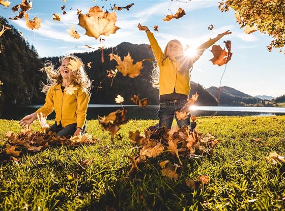 Zwei Kinder in gelben Jackets spielen fröhlich im Herbstlaub. Sie werfen bunte Blätter in die Luft, während die Sonne im Hintergrund scheint.