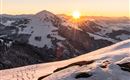 A picturesque mountain with snow-covered slopes and a warm sun on the horizon. The sky is clear and the landscape shines in soft colors.