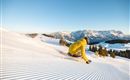 Ein Skifahrer trägt eine gelbe Jacke und fährt die perfekt präparierte Piste hinunter. Im Hintergrund sind schneebedeckte Berge und ein klarer blauer Himmel zu sehen.