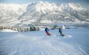 Two skiers are skiing over a snow-covered slope. In the background are majestic mountains and a clear sky.