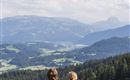 A couple is sitting on a bench, looking at a picturesque mountain landscape. The sky is clear and nature is lush green.
