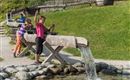 Children are playing at a water play area and experimenting with water. In the background, green meadows and wooden structures can be seen.