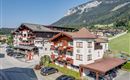 A picturesque Alpine landscape with several buildings in the foreground. The surroundings are green and mountainous, ideal for vacationers.