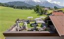 A rooftop terrace with sun loungers and umbrellas offers a beautiful view of green meadows and the mountains in the background. The environment is quiet and inviting.