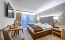 A modern hotel room with a double bed and a cozy chair. Large windows provide plenty of natural light and offer a view outside.