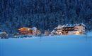 A wintry landscape with snow-covered hills. Cozy houses gently glow in the twilight.