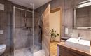 A modern bathroom with a glass shower and a wooden sink. The walls are in concrete gray, and in the background, there is a plant.