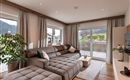 A modern living room with a comfortable gray couch and large windows. The room is bright and welcoming, with a view of a green landscape.