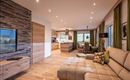 A modern living room with a comfortable sofa and wood-paneled walls. The open kitchen is bright and inviting, with large windows and plenty of daylight.