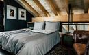 Ein stilvolles Schlafzimmer mit einem großen Bett und einem modernen Braun-Lederstuhl. Die Holzbalkendecke und die Bilder an der Wand verleihen dem Raum Wärme und Gemütlichkeit.