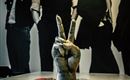 Eine Hand mit dem Peace-Zeichen steht auf einem Tisch. Im Hintergrund sind Silhouetten von vier Männern zu sehen.