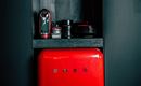 A red refrigerator stands in a modern cabinet. Above it are stylish dishes and a coffee machine.