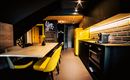 A modern kitchen with black cabinets and a yellow refrigerator. The dining area is equipped with a table and yellow chairs.