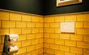 Ein modernes Badezimmer mit gelben Kacheln und schwarzer Wandfarbe. An der Wand hängt ein Bild, und es gibt eine Toilette mit Papierhaltern.