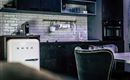 A modern kitchen with a stylish SMEG refrigerator and elegant black furniture. The walls are clad with white tiles, creating a minimalist look.