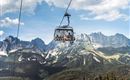 A cable car glides over green meadows and majestic mountains. The sky is clear and the landscape is stunning.