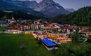 A picturesque landscape with a hotel complex, surrounded by mountains and forests. The outdoor area features a pool that is illuminated by lights.