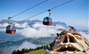 A cable car glides over an impressive mountain landscape. Two people are climbing on a large artwork in the shape of a face.