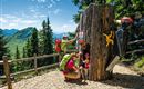 A wooden sculpture in the shape of a tree with a colorful bird and an entrance for playing. Two hikers, an adult and a child, are happily interacting with the sculpture in the woods.