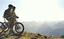 Two cyclists are standing on a high trail, admiring the mountain landscape. The sun is shining over the peaks, providing a breathtaking view.