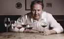 A man is sitting at a table with several wine glasses in front of him. He is holding a glass of red wine and looking straight into the camera.