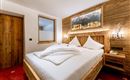 A cozy bedroom with a wooden bed and white pillows. A landscape picture hangs on the wall, and the floor is covered with a red carpet.