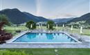 A beautiful pool with sun loungers, surrounded by green meadows and mountains. The sky is clear and shows soft colors of the sunset.