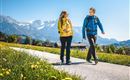 A couple is hiking on a picturesque path in the mountains. The sunny landscape is surrounded by green meadows and snow-covered peaks.