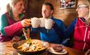 Three friends toast with mugs and enjoy a meal in a cozy setting. On the table, there's a plate with delicious treats.