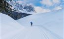 A skier is running on a snowy track. In the background, majestic mountains and a blue sky can be seen.