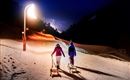 Two people are pulling sleds on a lit snow slope at night. In the background, the lights of the mountain are shining.