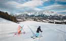 Two skiers are skiing on a snowy slope. In the background, impressive mountains and a blue sky are visible.