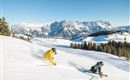 Two skiers are skiing over fresh snow in the mountains. In the background, majestic mountains and a clear blue sky can be seen.