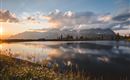A tranquil lake reflects the mountains and the sky at sunset. The edge is lined with green meadows and flowers.
