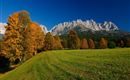 A picturesque landscape with colorful autumn trees and majestic mountains in the background. The sky is clear and blue, while a green meadow characterizes the foreground.
