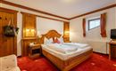 A cozy bedroom with a large double bed and wooden paneling. The room has red carpets and windows with decorative curtains.