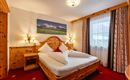 A cozy bedroom with a wooden bed and red carpet. The room is brightly lit and has large windows looking outside.