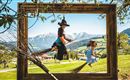 A girl and a witch are happily flying on brooms in a picturesque landscape frame. In the background, green hills and mountains are visible.