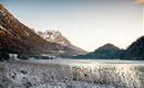A picturesque winter landscape with snow-covered mountains and a frozen lake. In the foreground, tall grasses grow along the shore.