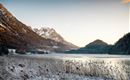 A winter landscape with a frozen lake and snow-covered mountains in the background. The sky is clear and the sun is shining gently.