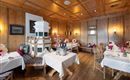 A cozy restaurant with wooden walls and elegantly set tables. In the center stands a traditional tiled stove.