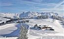 A winter landscape with snow-covered mountains and a small house. The sky is clear and blue.