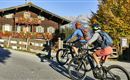 Two cyclists are riding on a path near a traditional wooden house. In the background, mountains and colorful trees can be seen.