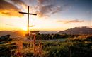A cross silhouette against a stunning sunset over the mountains. Green meadows and flowers complement the peaceful scene.